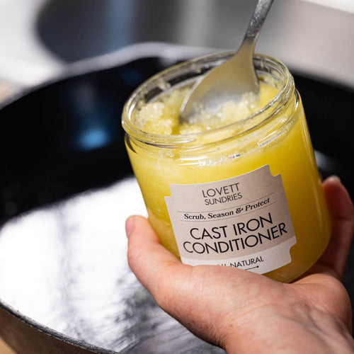 Hand holding a jar of 'Cast Iron Conditioner' with a spoon inside, labeled 'Lovett Sundries'.